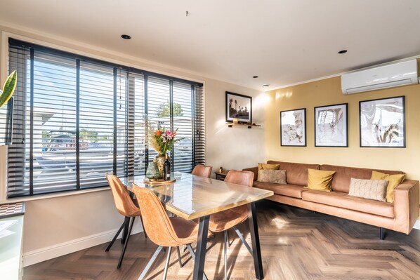 A modern living room with a glass dining table, leather sofa, and large windows with blinds. - Unieke en Moderne Water Villa in de Buurt van Schiphol en Amsterdam (Aalsmeer)