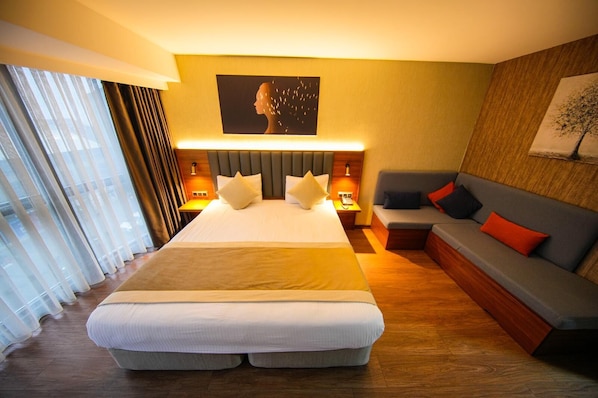 A hotel room with a large bed, a sofa, a wall-mounted painting, and a window with curtains.
