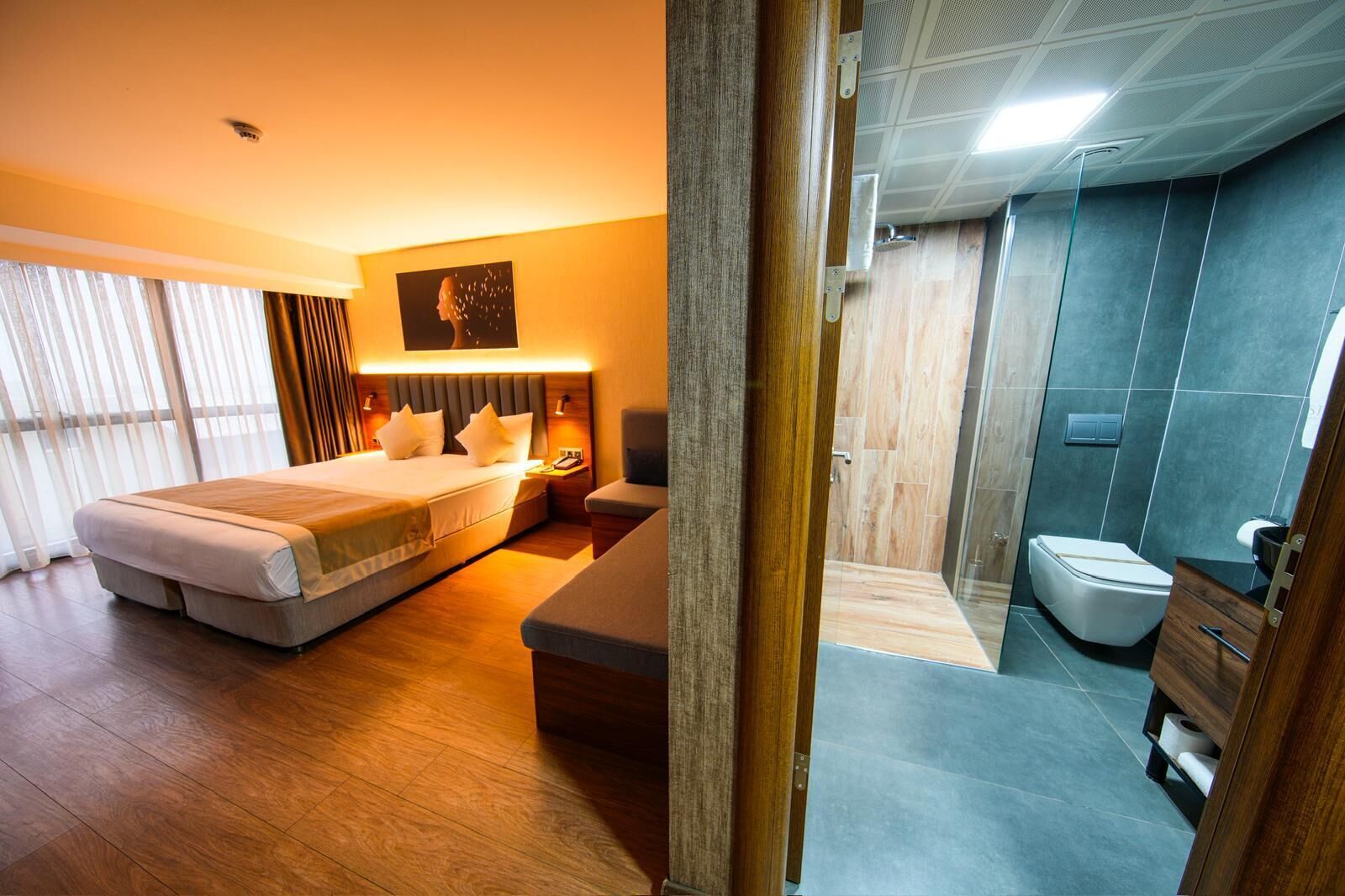 A hotel room with a large bed, bedside tables, a nightstand, and a wall-mounted artwork. The bathroom features a shower, toilet, and a wooden vanity.