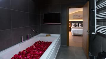 Un baño moderno con una bañera llena de pétalos de rosa, un televisor montado y una habitación visible con una cama y un cuadro en la pared.