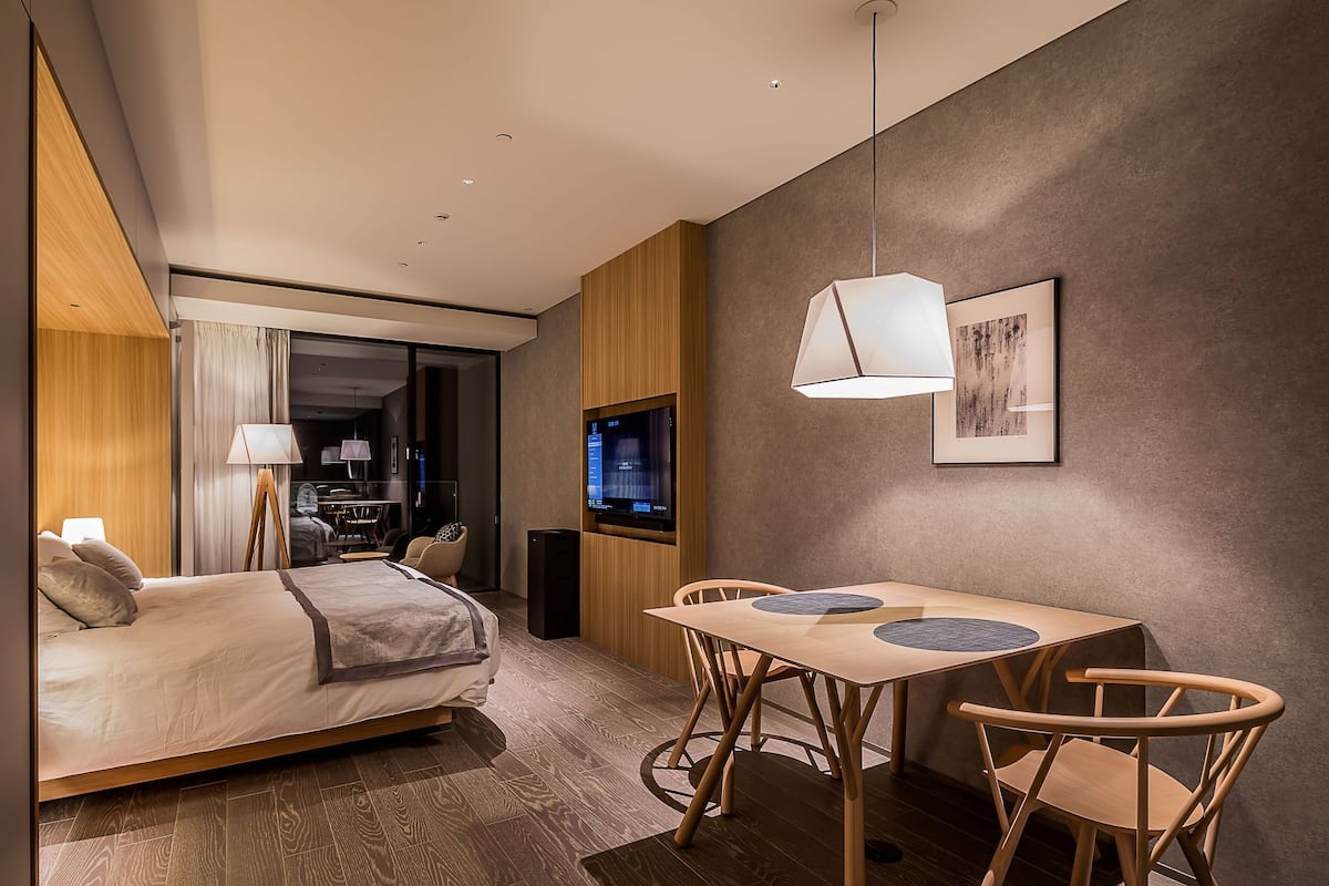 Una habitación de hotel moderna con cama, una mesa de comedor con sillas, televisión e iluminación colgante.