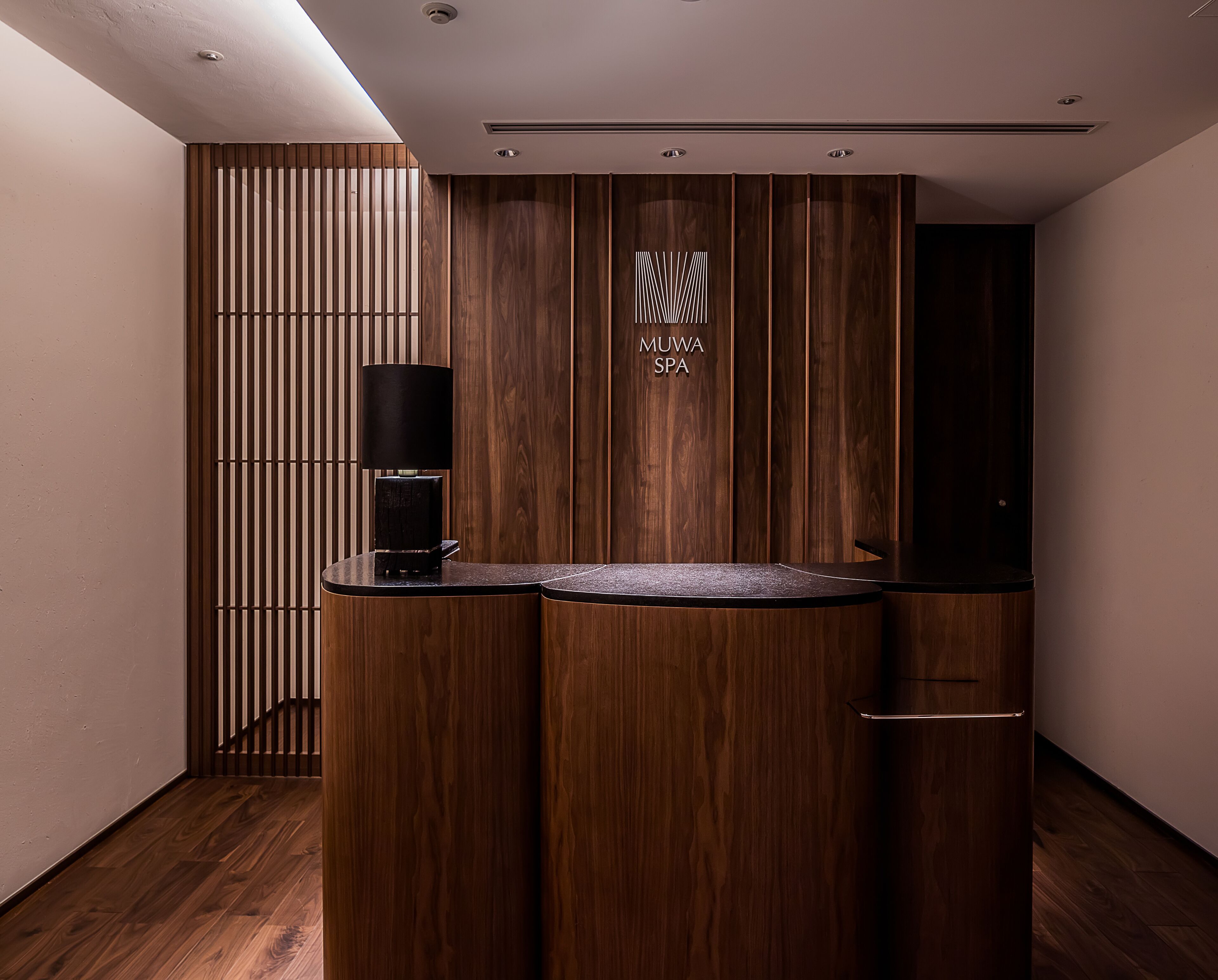 A reception area with a wooden reception desk, a black lamp, and a vertical wooden panel with the text "MUWA SPA".