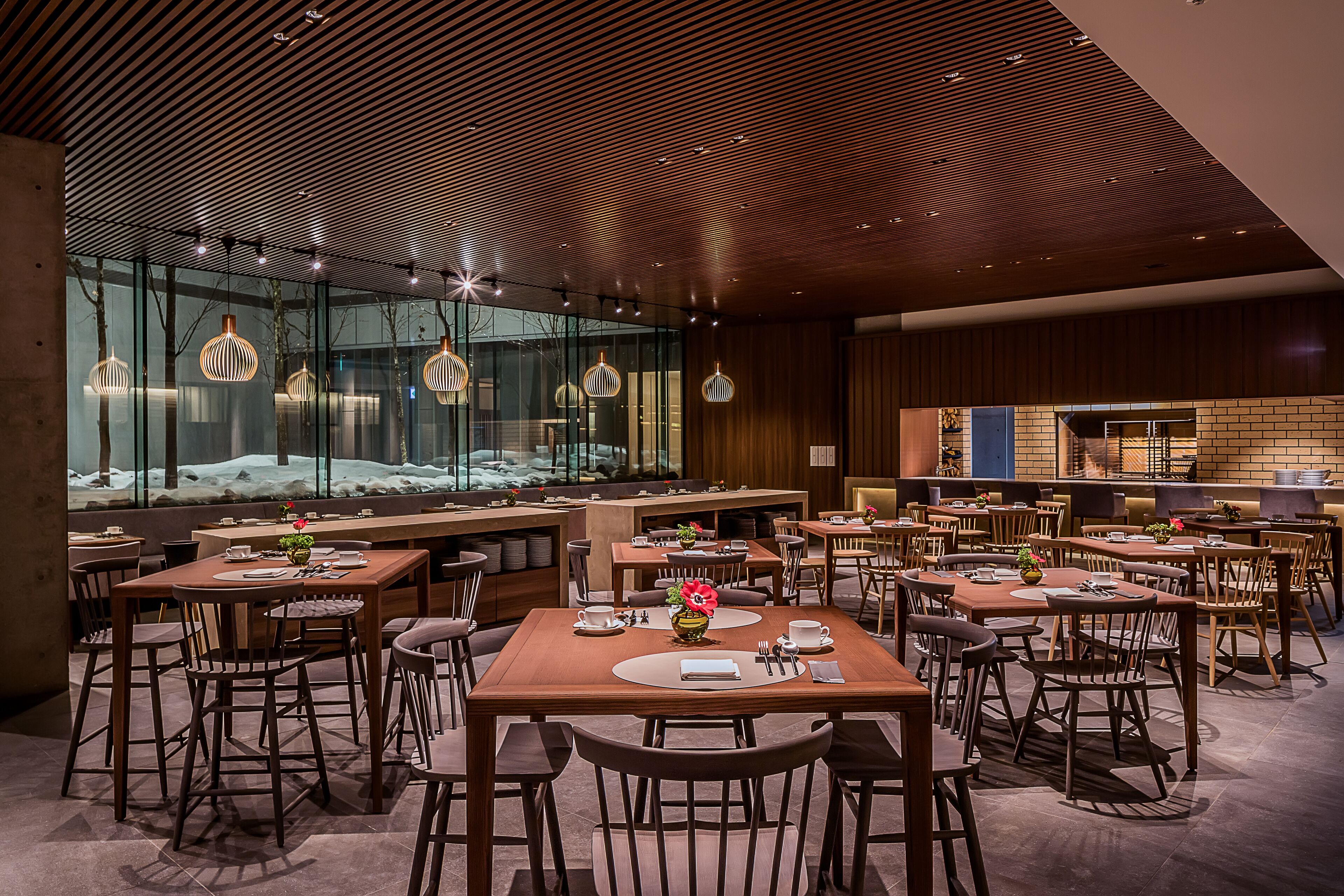 Un restaurante moderno con mesas y sillas de madera, ventanas amplias y lámparas colgantes.