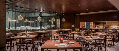Un restaurante moderno con mesas y sillas de madera, ventanas amplias y lámparas colgantes.