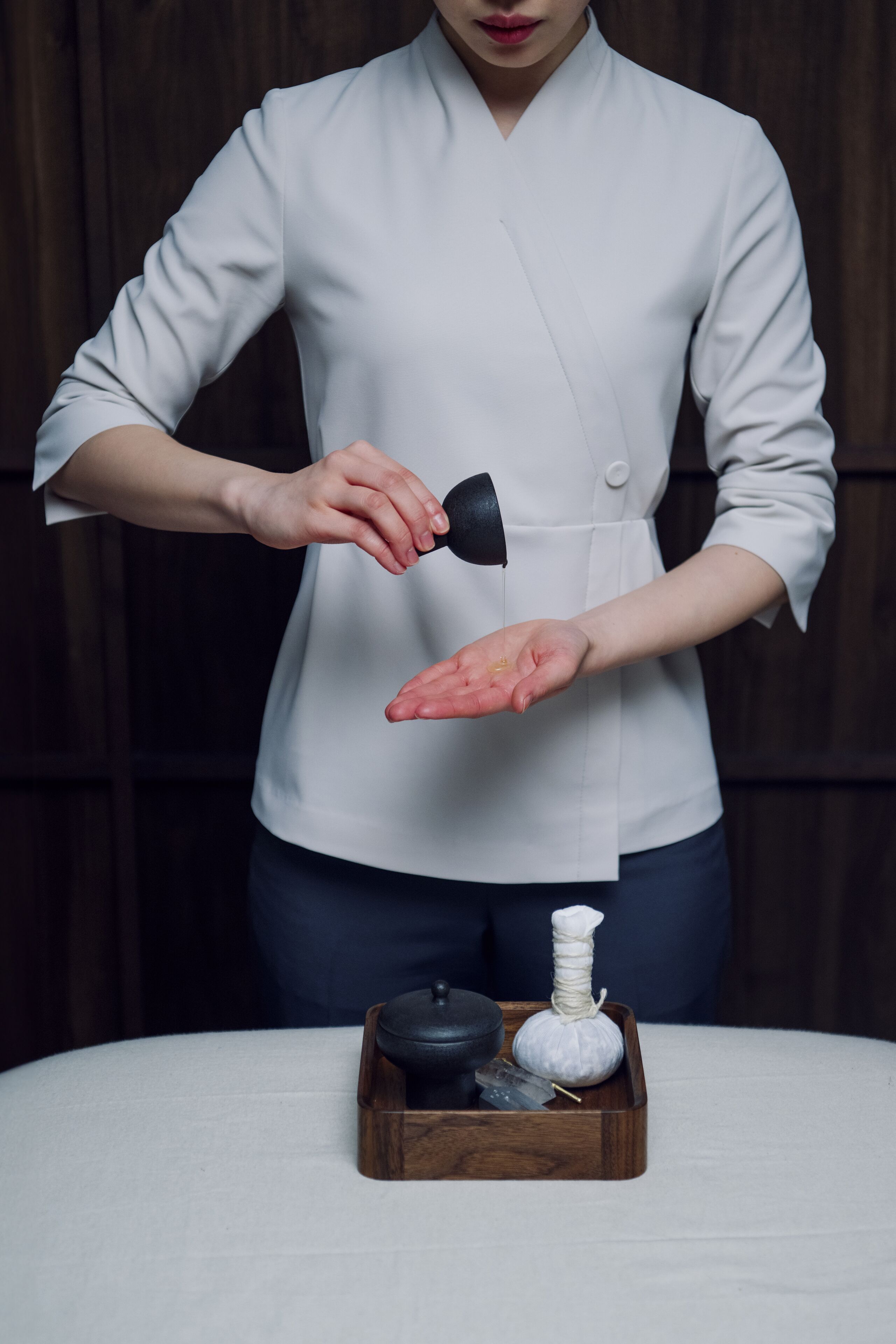 A person in a white coat holding a black object, standing next to a wooden tray with a white object and a black object on a white table.