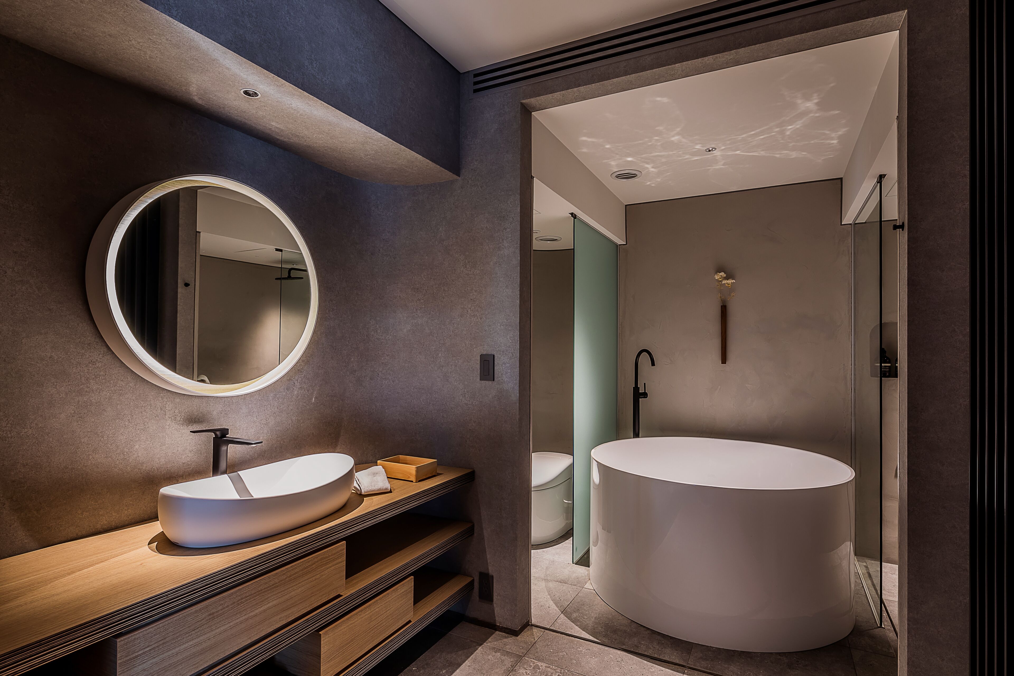 A modern bathroom with a round mirror, a wooden vanity, a large white sink, and a freestanding bathtub.