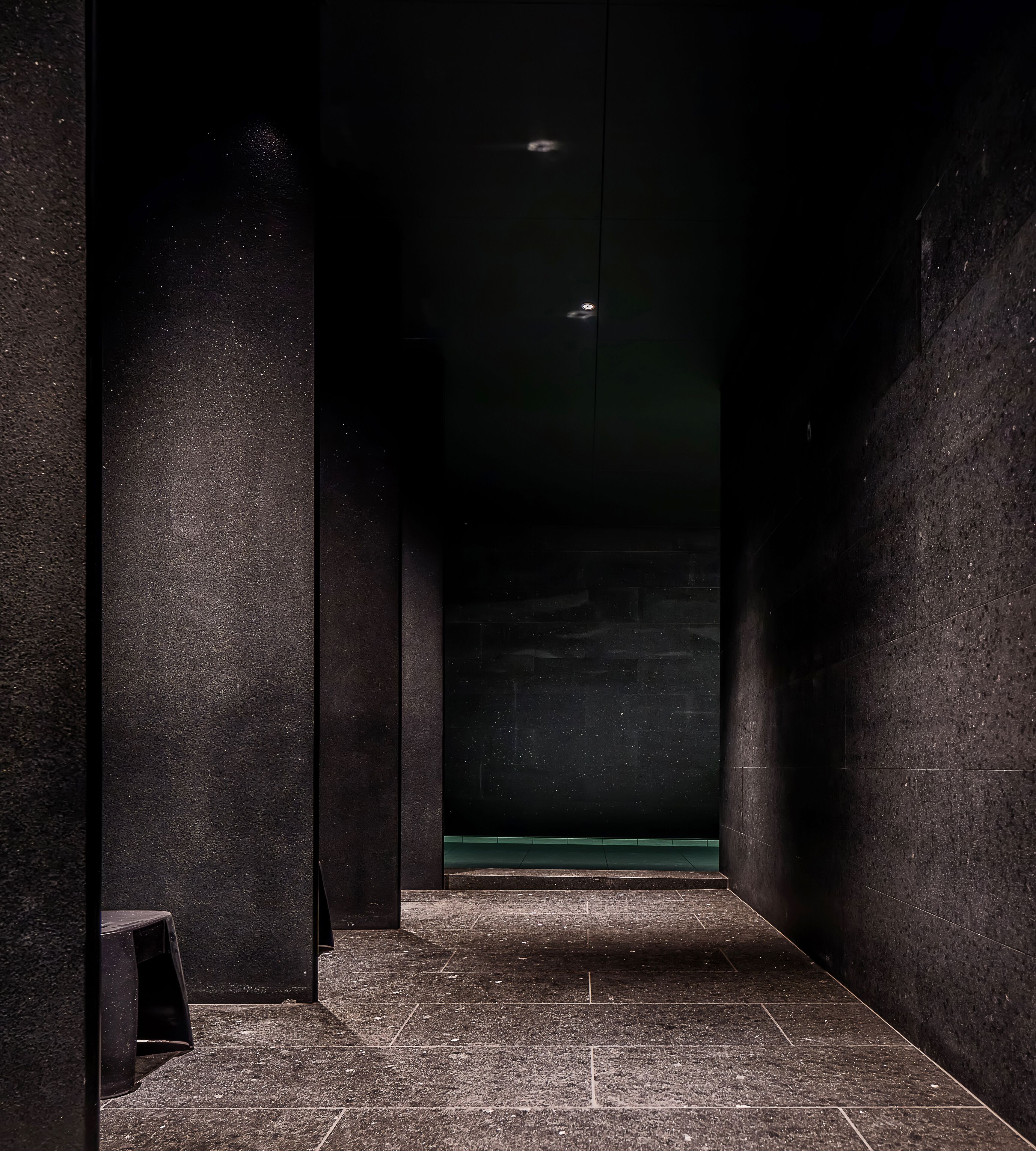 A narrow hallway with dark walls and a tiled floor, leading to a green-lit area.