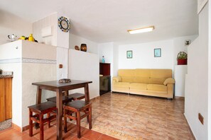 A living room with a sofa, a dining table with chairs, a kitchen area with a refrigerator, and a tiled floor.