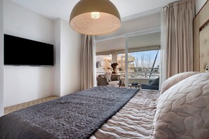 A hotel room with a bed, a flat-screen TV, a large window, and a balcony with a view of boats.