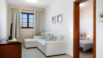 Una habitación con un sofá blanco, un escritorio de madera, una ventana con cortinas y una cama con colcha estampada.