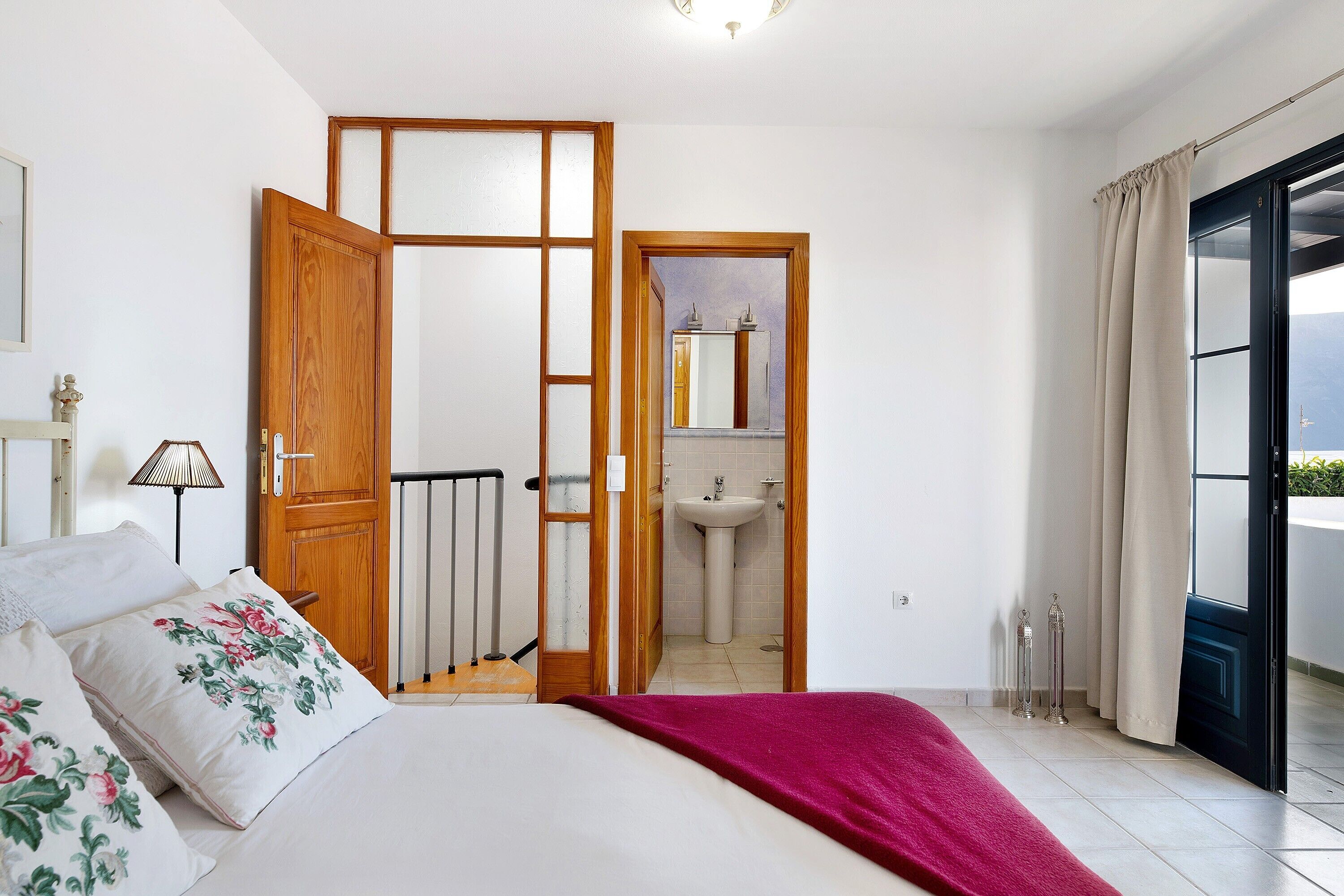 Een slaapkamer met een bed, een houten deur, een badkamer met een wastafel en uitzicht door een raam.