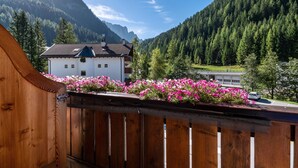 A wooden balcony with a view of a white building with a dark roof, surrounded by vibrant pink flowers and a forested mountain backdrop. - Apartment "Stoffelapartments - Marie" with Mountain View, Shared Terrace & Wi-Fi (Wolkenstein)