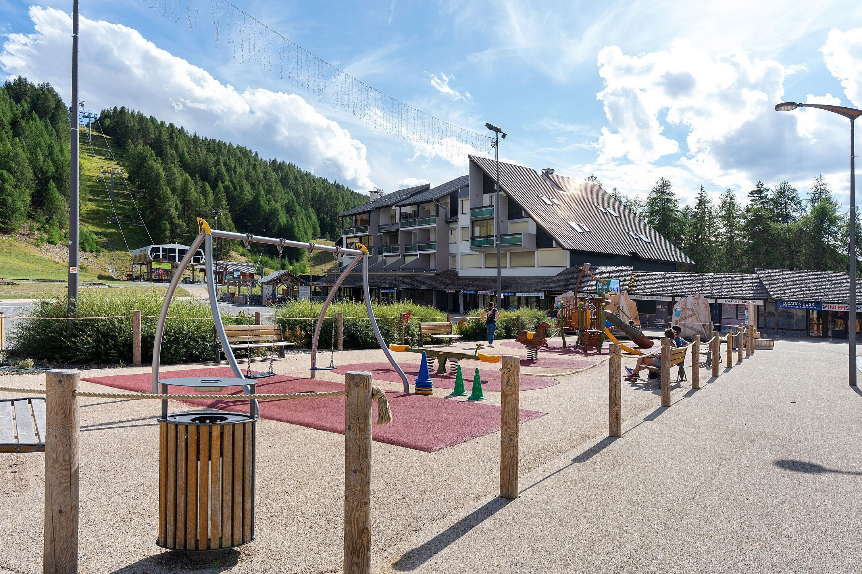 Un terrain de jeux avec des balançoires et des toboggans, un banc en bois et, au fond, un bâtiment doté de balcons.