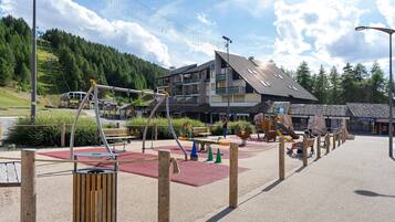 Un terrain de jeux avec des balançoires et des toboggans, un banc en bois et, au fond, un bâtiment doté de balcons.