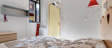 A bedroom with a bed, a window, a door, and a wall-mounted lamp.