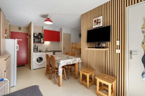 A compact living space with a kitchenette, a dining area with a table and chairs, a washing machine, and a television mounted on a wooden wall. - Apartment 'Le Relais Du Devoluy' with Mountain View and Terrace (Le Dévoluy)
