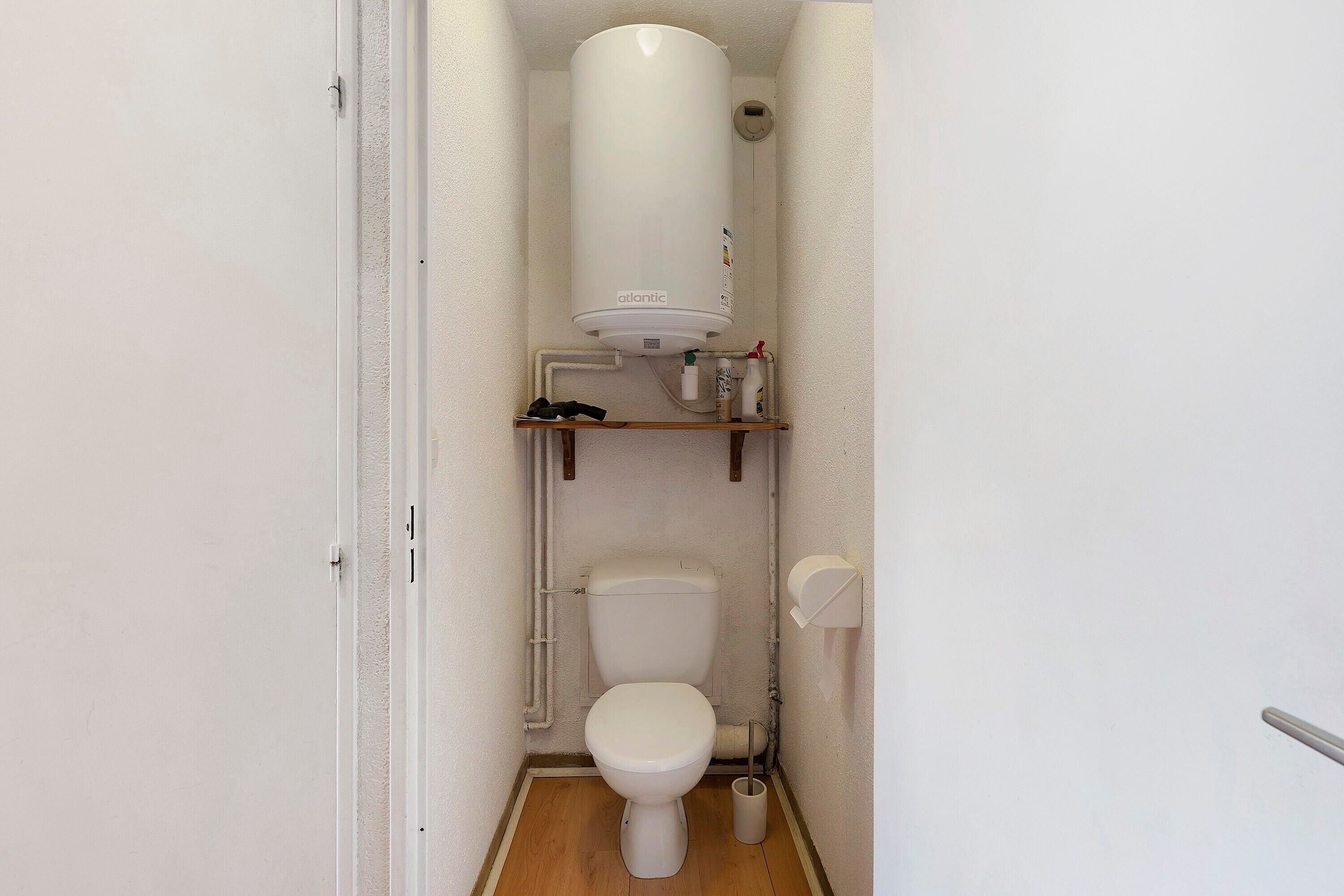 A small, compact bathroom with a toilet, a white pedestal heater, and a shelf with toiletries.