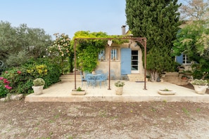 Une terrasse couverte avec une table et des chaises, une pergola et une maison avec une porte et des fenêtres bleues.