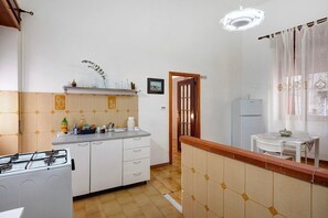 Une cuisine équipée d’une cuisinière à gaz, de meubles blancs, d’une petite étagère avec des bouteilles et d’une fenêtre avec un rideau.