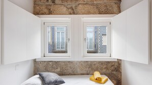A bedroom with a stone wall, two windows with patterned curtains, a bed with white bedding, a pillow, and a rolled towel. - Holiday Home 'Lidador House' with Private Terrace, Private Garden and Wi-Fi (Vila do Conde)