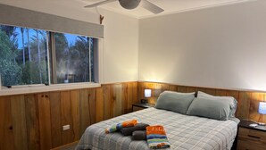 Un dormitorio con cama, paneles de madera, ventana con vista a árboles, mesita de noche con lámpara y ventilador de techo.