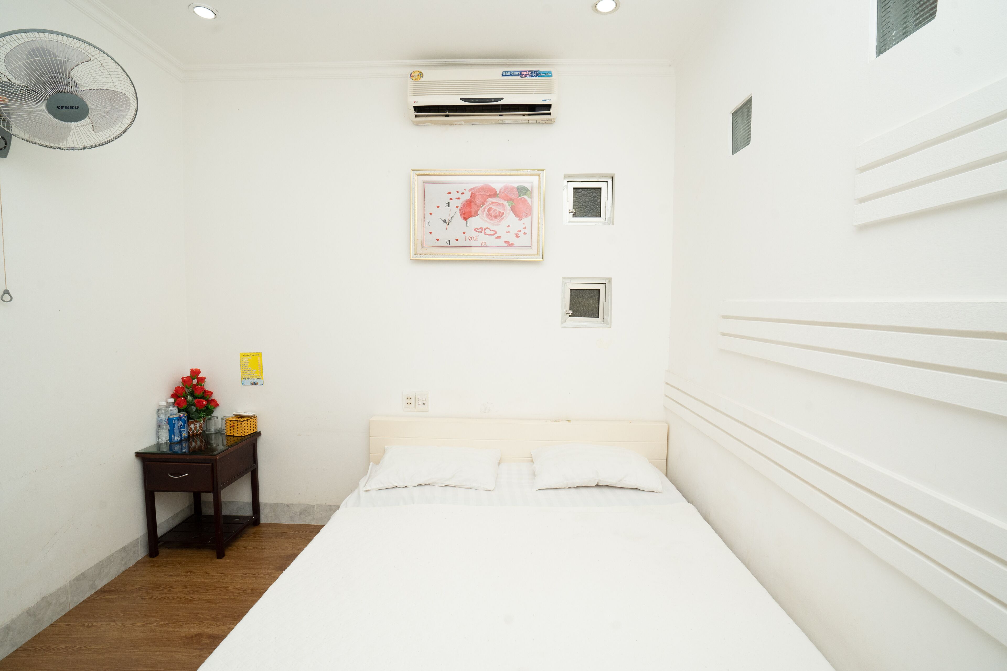 A clean, white room with a bed, a small bedside table, a fan, an air conditioner, and a framed picture on the wall.