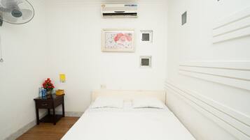 A clean, white room with a bed, a small bedside table, a fan, an air conditioner, and a framed picture on the wall.