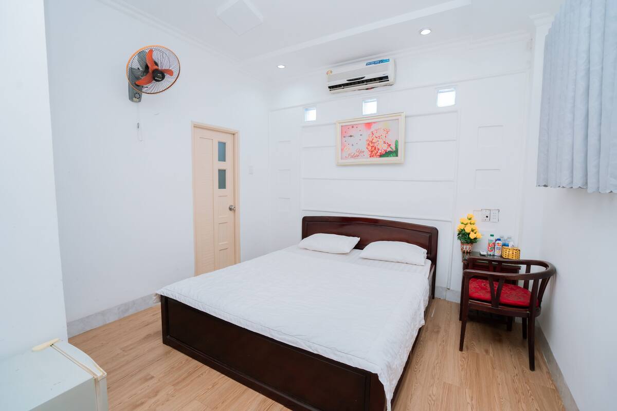 A bedroom with a wooden bed, a wall-mounted fan, an air conditioner, a door, and a small table with a chair.