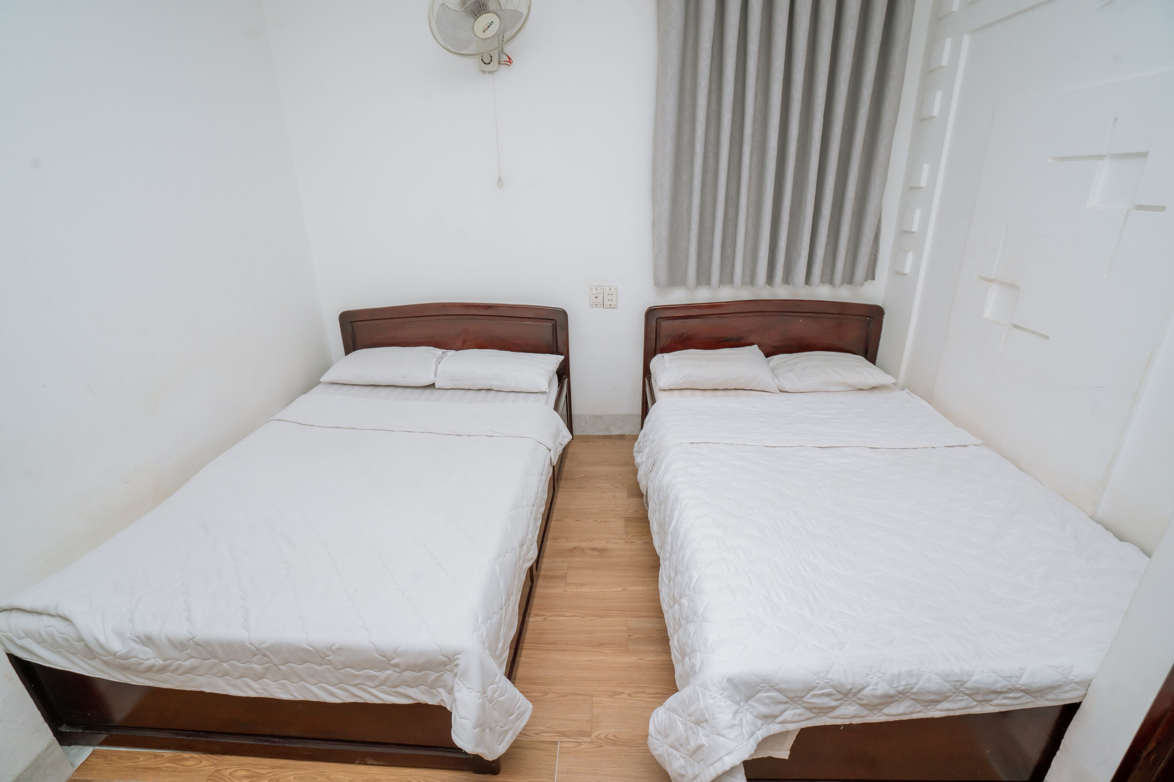 Une chambre avec deux lits simples, un ventilateur de plafond, une fenêtre avec des rideaux et un plancher en bois.