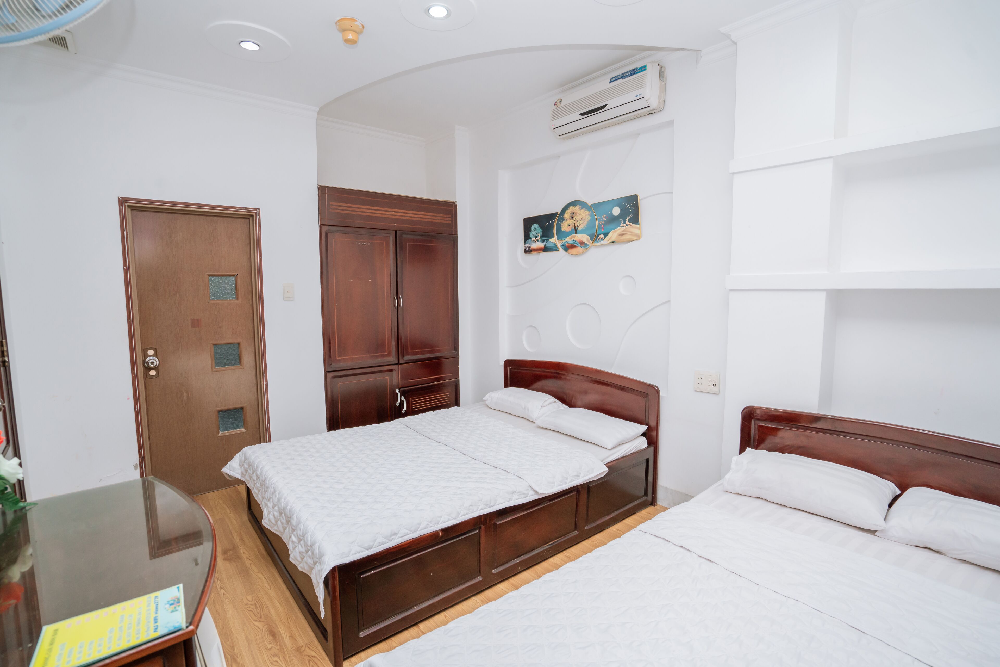 Een hotelkamer met een houten bed, een kledingkast van hout, een klein tafeltje met een stoel, en een luchtverkoeler aan de muur.