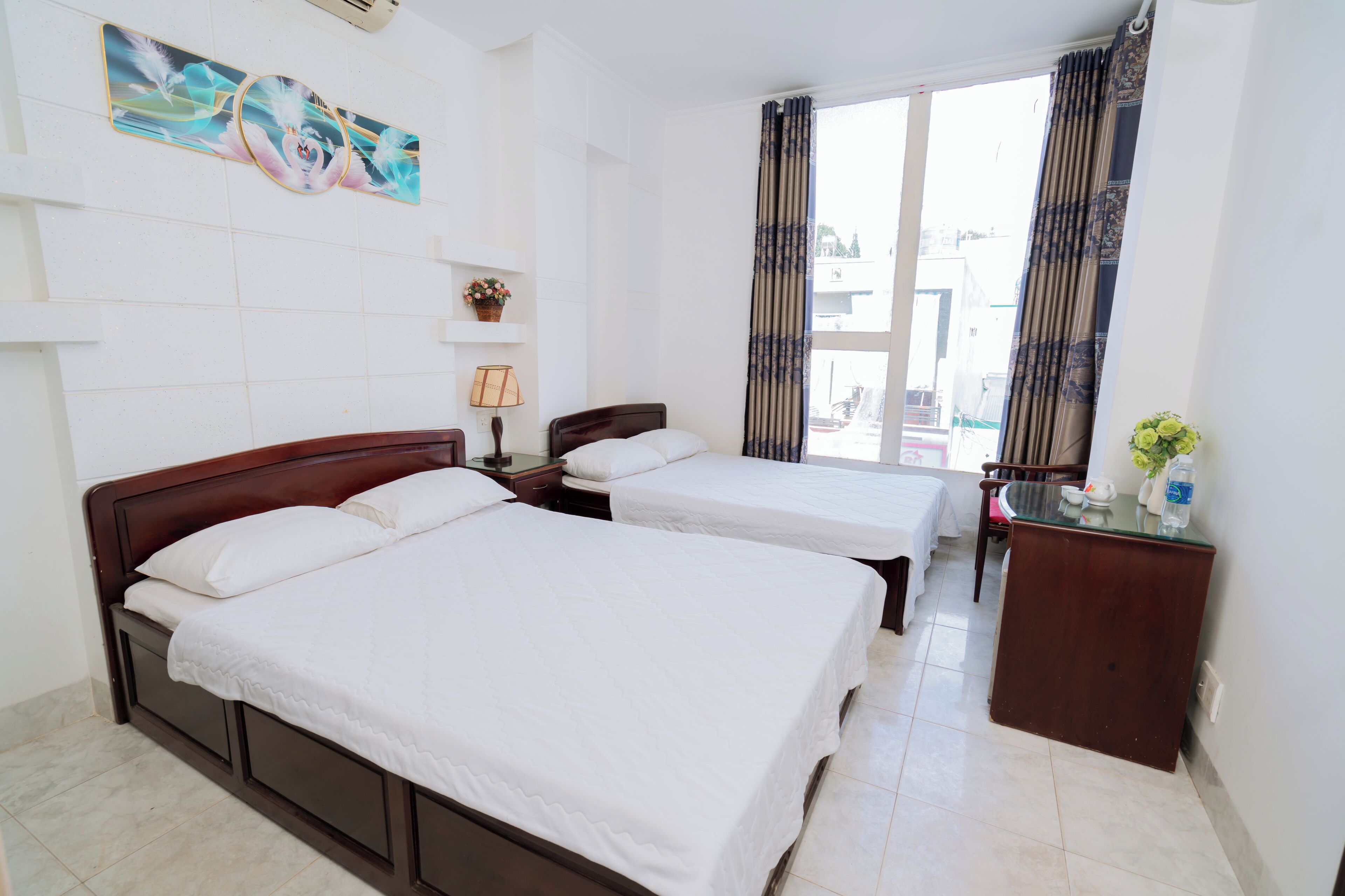 A hotel room with two beds, a desk, a chair, a window with curtains, a wall-mounted shelf with a plant, and a painting.