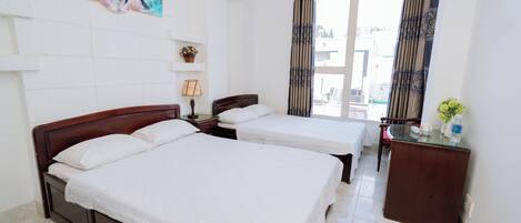 A hotel room with two beds, a desk, a chair, a window with curtains, a wall-mounted shelf with a plant, and a painting.