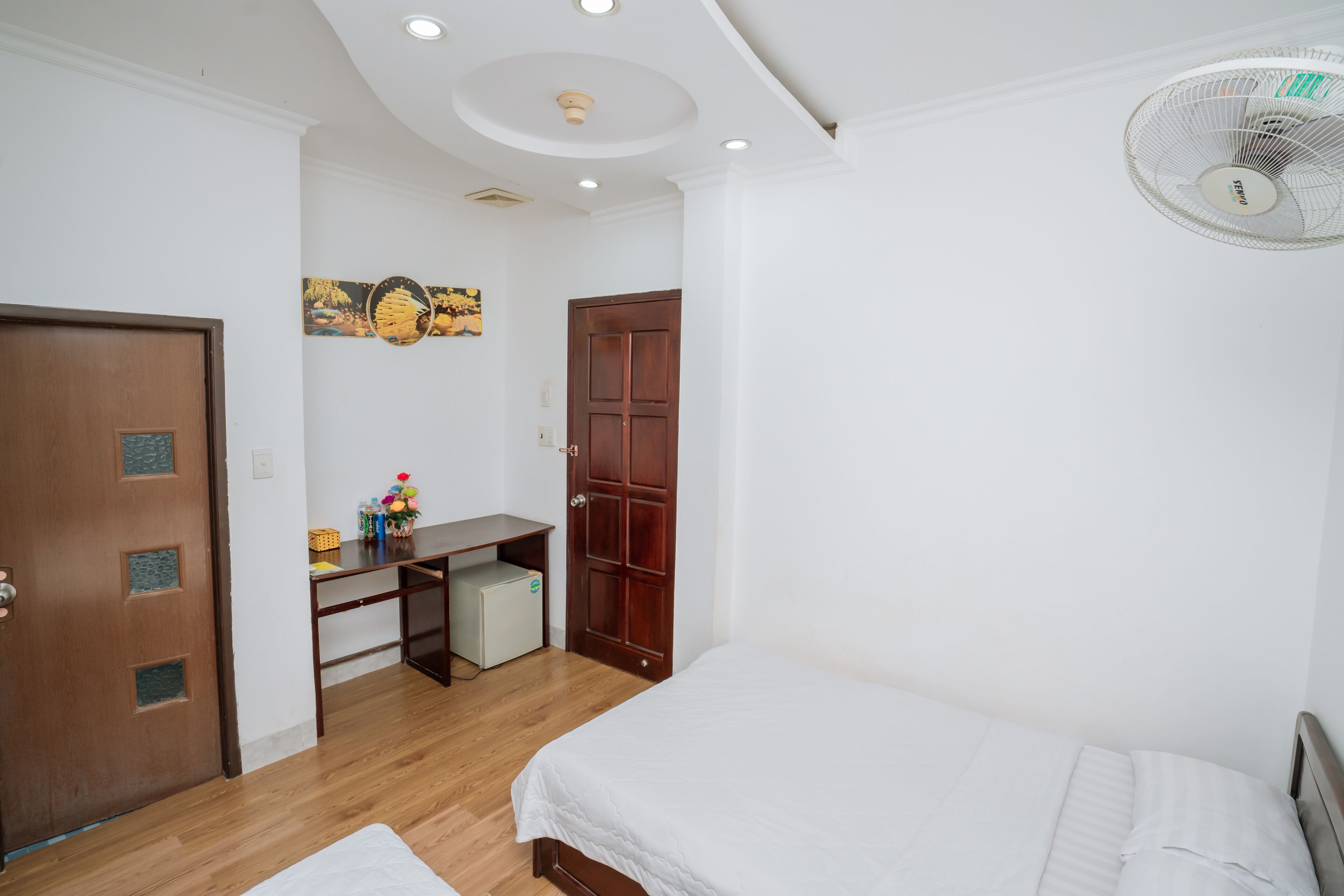 A bedroom with a bed, a desk, a small fridge, and a ceiling fan.