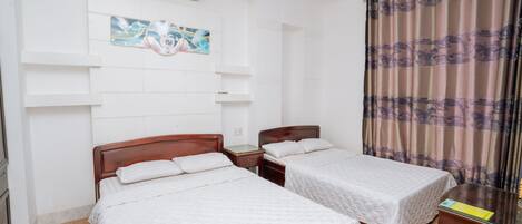 A hotel room with two single beds, a wooden headboard, a bedside table, a nightstand, a wall-mounted air conditioner, and a window with patterned curtains.