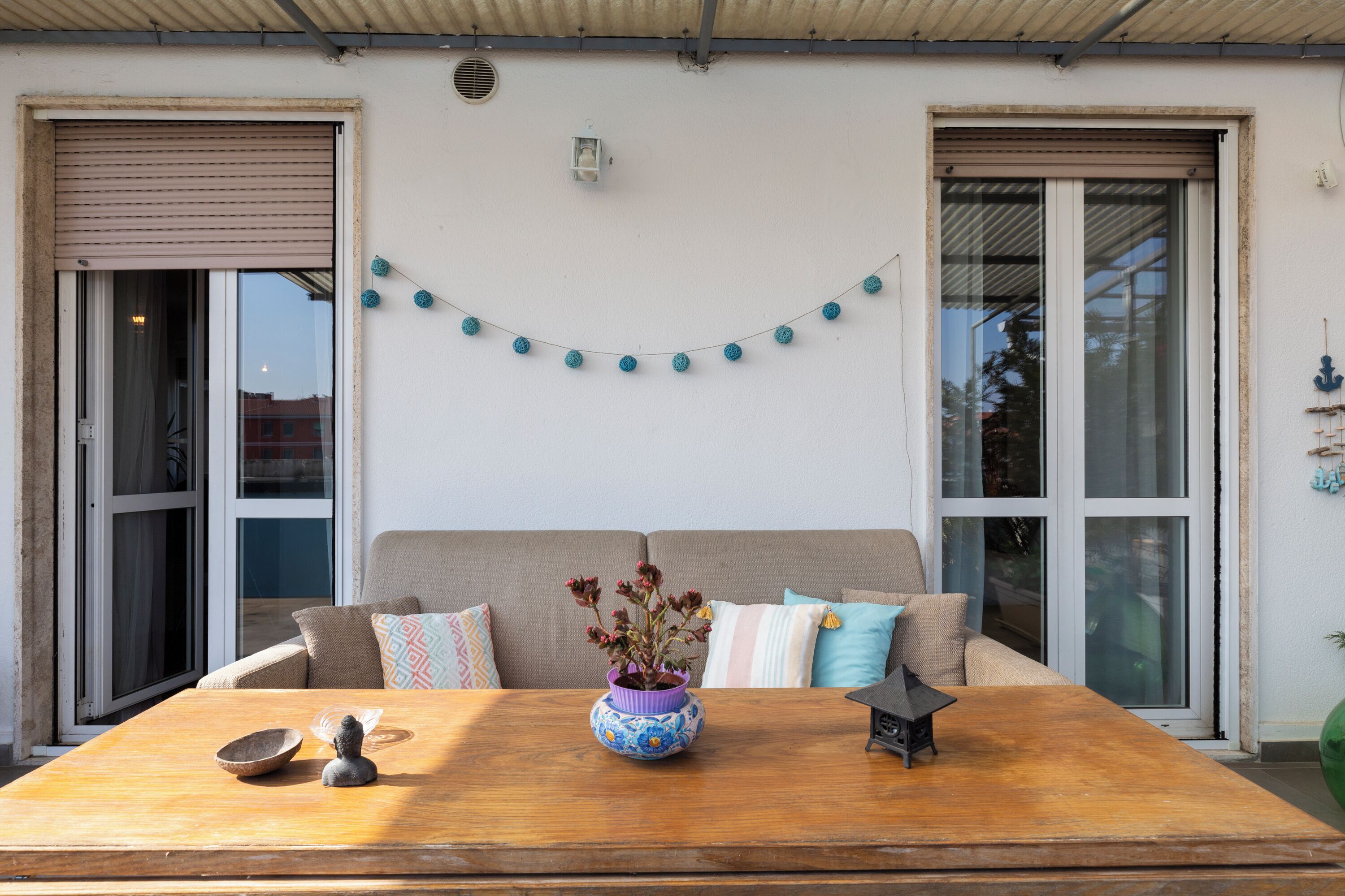 A room with a wooden table, a sofa with cushions, a potted plant, and a string of decorative balls on the wall.