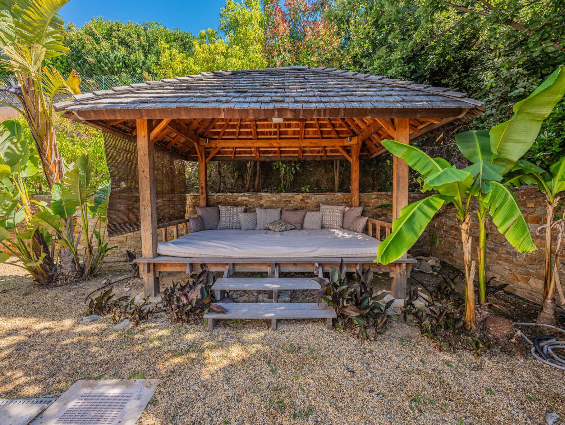 A wooden gazebo with a bench and steps, surrounded by tropical plants and a stone wall.