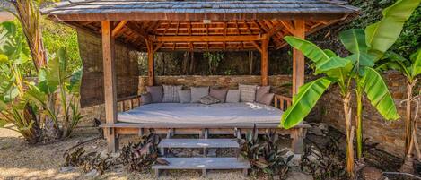 A wooden gazebo with a bench and steps, surrounded by tropical plants and a stone wall.