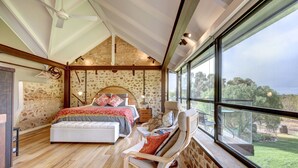 Un dormitorio con una cama grande, una zona de estar con una silla y vistas al exterior a través de amplias ventanas.
