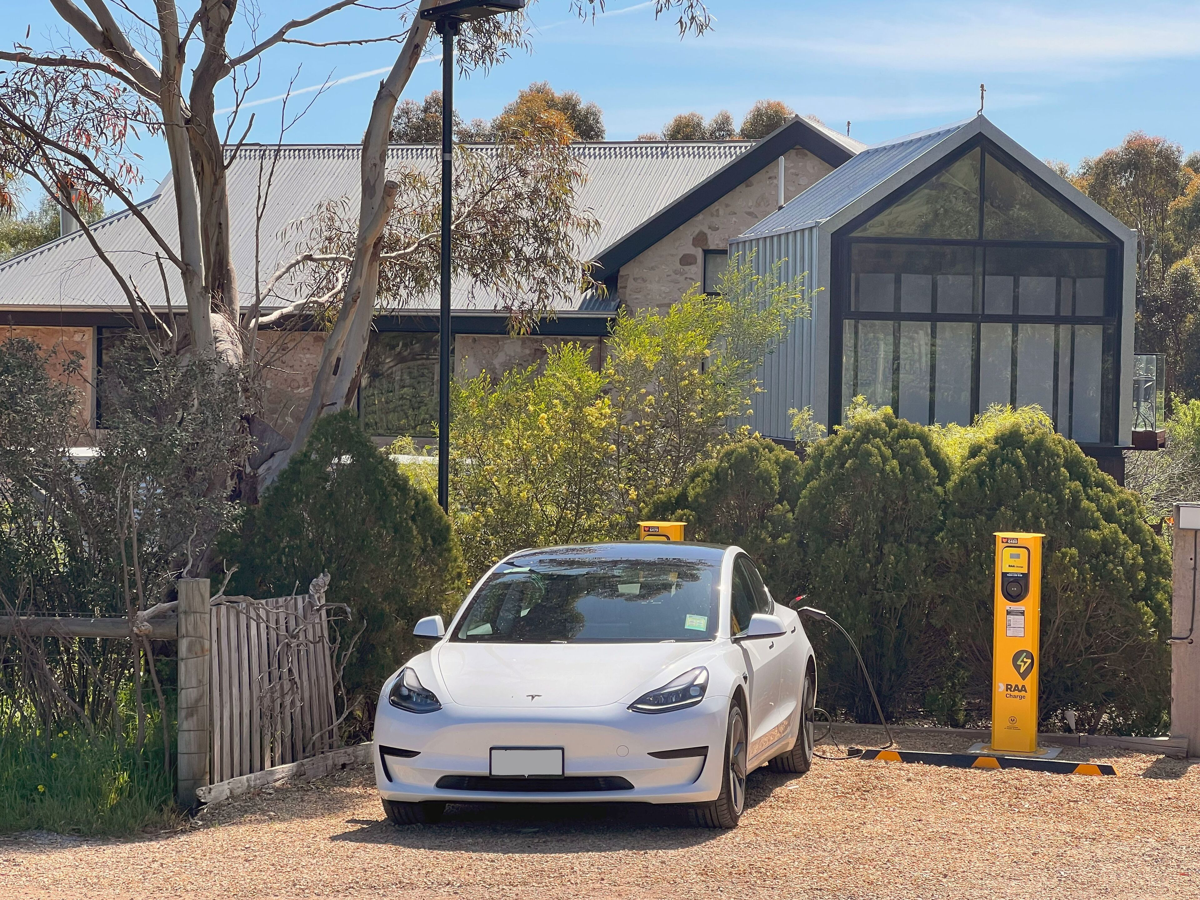 A white car parked at an electric vehicle charging station, with a house and trees in the background.