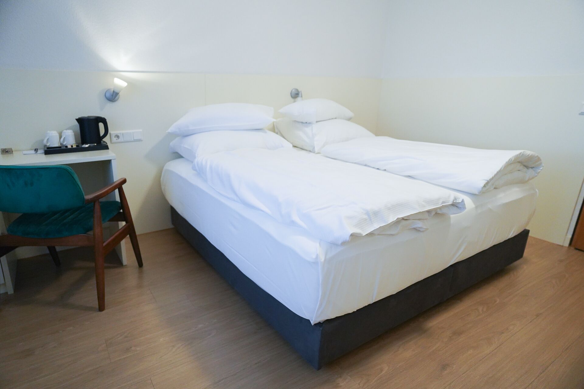 A neatly made bed with white linens, a wooden chair, a small table with a coffee maker, and a wall-mounted light.