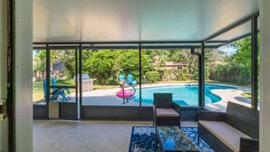 A glass-enclosed patio with a pool, outdoor furniture, and a view of the surrounding greenery.