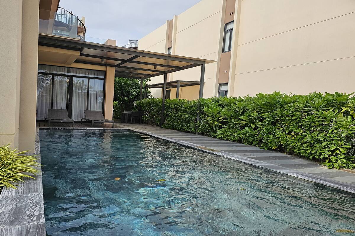 Une piscine dotée d’une enceinte en verre, située à côté d’un bâtiment avec un patio couvert et offrant une vue sur un quartier résidentiel.
