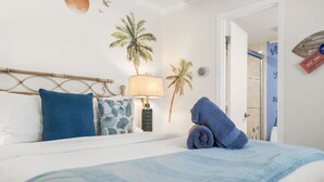Una cama bien hecha con una manta azul y almohadas, una lámpara de pared y un adhesivo de pared con motivos tropicales.
