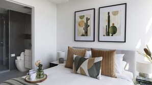 Un dormitorio moderno con cama, almohadas, una mesita de noche, una obra de arte enmarcada y un baño visible a través de una puerta abierta.