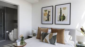 A modern bedroom with a bed, pillows, a bedside table, a framed artwork, and a bathroom visible through an open door.