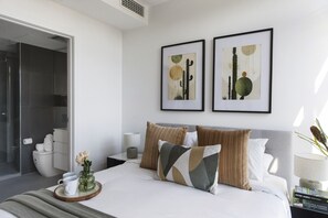 Un dormitorio moderno con cama, almohadas, una mesita de noche, una obra de arte enmarcada y un baño visible a través de una puerta abierta.