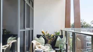 Een balkon met een eettafel gedekt voor twee personen, een bank met groene, gestreepte kussens, en uitzicht op de stad.