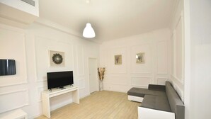 Een woonkamer met een witte tv-meubel, een flatscreen televisie, een grijze bank en een witte bankje.