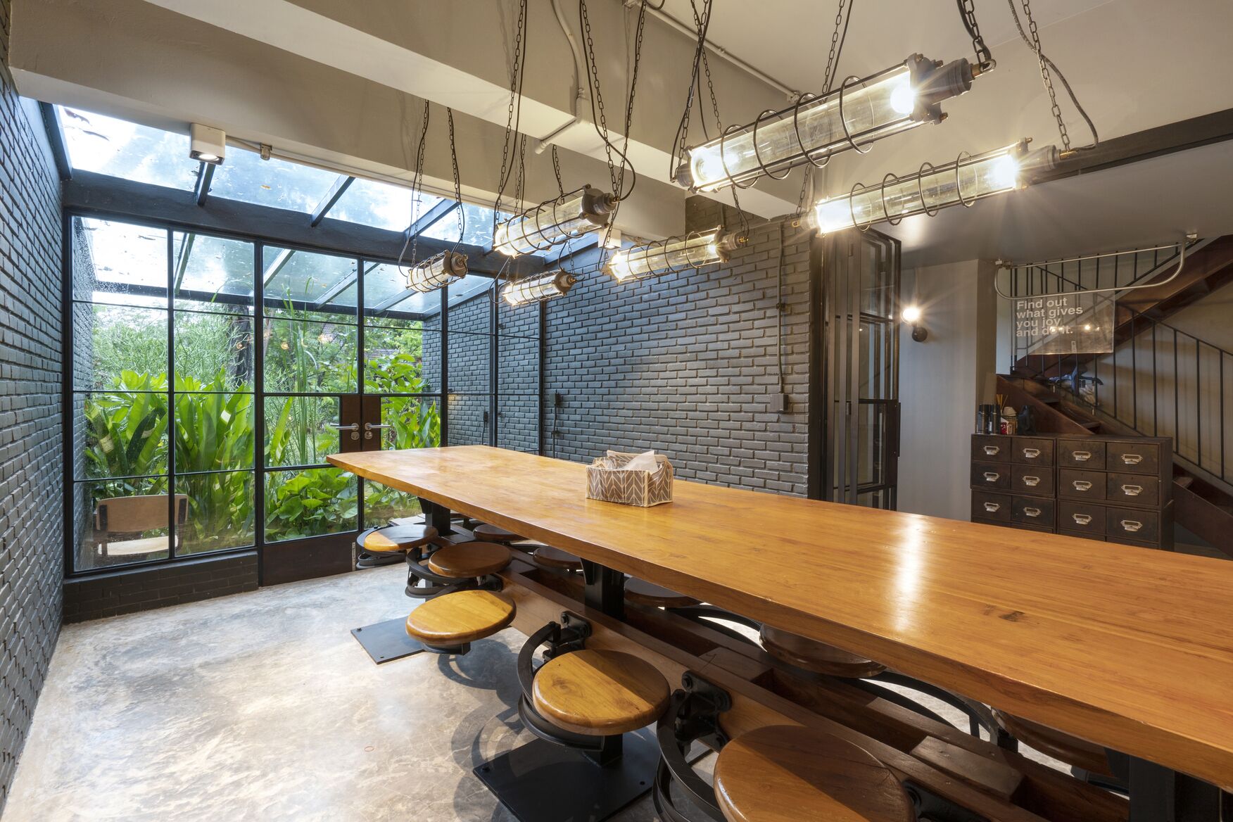 A modern dining area with a long wooden table, multiple stools, and a view through large windows to a green outdoor space.