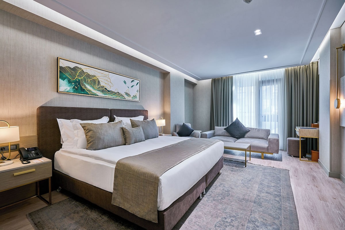 A modern hotel room with a large bed, a desk with a lamp, a sofa, and a large window with curtains.